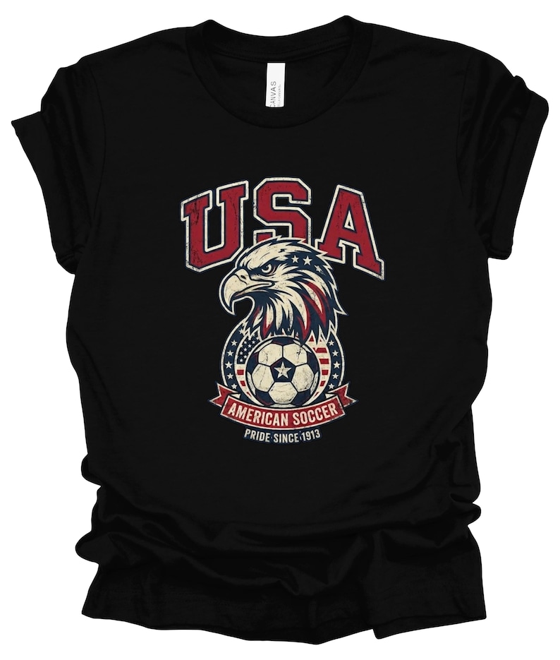 K&ouml;nnte beinhalten: Ein schwarzes Kurzarm-T-Shirt mit einer Grafik eines Wei&szlig;kopfseeadler-Kopfes &uuml;ber einem Fu&szlig;ball, mit dem Text USA, AMERICAN SOCCER und PRIDE SINCE 1913. Das Design enth&auml;lt rote, wei&szlig;e und blaue Farben mit einer abgenutzten, vintage-artigen Textur.