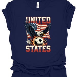 Op de afbeelding: Een marineblauw t-shirt met korte mouwen en een afbeelding van een Amerikaanse zeearend met gespreide vleugels boven een voetbal omringd door vlammen, tegen een wapperende Amerikaanse vlag. De tekst UNITED STATES staat in dikke, rood-witte letters.