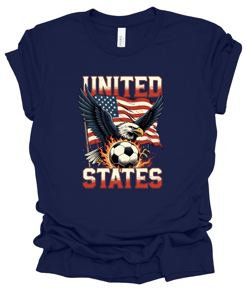 Op de afbeelding: Een marineblauw t-shirt met korte mouwen en een afbeelding van een Amerikaanse zeearend met gespreide vleugels boven een voetbal omringd door vlammen, tegen een wapperende Amerikaanse vlag. De tekst UNITED STATES staat in dikke, rood-witte letters.