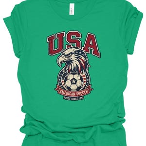 K&ouml;nnte beinhalten: Ein hellgr&uuml;nes Kurzarm-T-Shirt mit einer Grafik eines Wei&szlig;kopfseeadler-Kopfes &uuml;ber einem Fu&szlig;ball. Der Text lautet USA, AMERICAN SOCCER und PRIDE SINCE 1913. Das Design enth&auml;lt rote, wei&szlig;e und blaue Farben mit einer abgenutzten Textur.