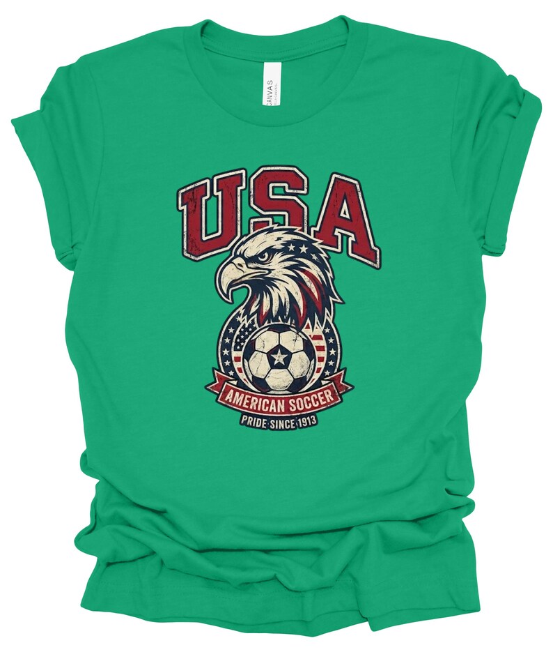 K&ouml;nnte beinhalten: Ein hellgr&uuml;nes Kurzarm-T-Shirt mit einer Grafik eines Wei&szlig;kopfseeadler-Kopfes &uuml;ber einem Fu&szlig;ball. Der Text lautet USA, AMERICAN SOCCER und PRIDE SINCE 1913. Das Design enth&auml;lt rote, wei&szlig;e und blaue Farben mit einer abgenutzten Textur.