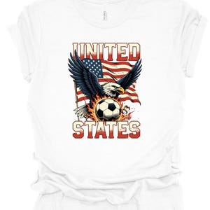 Op de afbeelding: Een wit t-shirt met een grafische print van een Amerikaanse zeearend met gespreide vleugels voor een wapperende Amerikaanse vlag. Onder de adelaar bevindt zich een voetbal, omringd door vlammen. De tekst UNITED STATES is in het rood gedrukt.