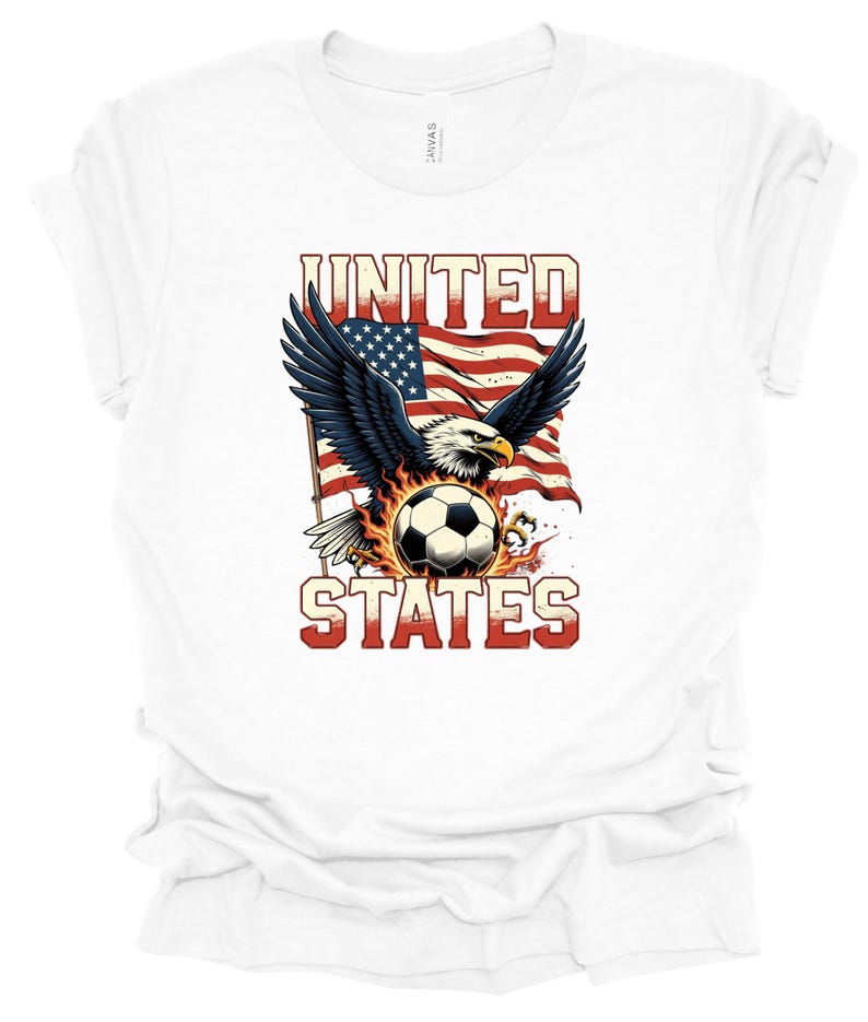 Op de afbeelding: Een wit t-shirt met een grafische print van een Amerikaanse zeearend met gespreide vleugels voor een wapperende Amerikaanse vlag. Onder de adelaar bevindt zich een voetbal, omringd door vlammen. De tekst UNITED STATES is in het rood gedrukt.