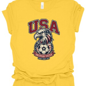 K&ouml;nnte beinhalten: Ein leuchtend gelbes Kurzarm-T-Shirt mit einer verwaschenen Grafik eines Wei&szlig;kopfseeadler-Kopfes &uuml;ber einem Fu&szlig;ball. Der Text lautet USA, AMERICAN SOCCER und PRIDE SINCE 1913. Das Design enth&auml;lt die Farben Rot, Wei&szlig; und Blau mit Sternen.