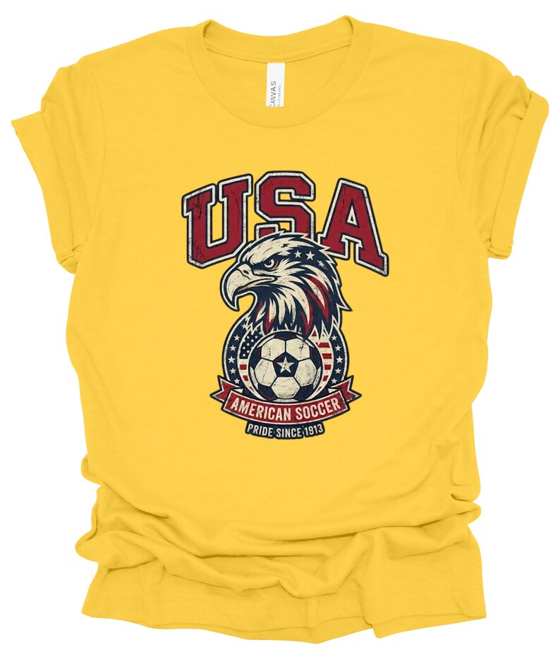K&ouml;nnte beinhalten: Ein leuchtend gelbes Kurzarm-T-Shirt mit einer verwaschenen Grafik eines Wei&szlig;kopfseeadler-Kopfes &uuml;ber einem Fu&szlig;ball. Der Text lautet USA, AMERICAN SOCCER und PRIDE SINCE 1913. Das Design enth&auml;lt die Farben Rot, Wei&szlig; und Blau mit Sternen.
