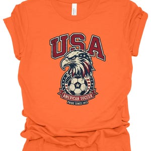K&ouml;nnte beinhalten: Ein orangefarbenes T-Shirt mit kurzen &Auml;rmeln und einer Grafik eines Wei&szlig;kopfseeadler-Kopfes &uuml;ber einem Fu&szlig;ball. Der Text lautet USA, AMERICAN SOCCER und PRIDE SINCE 1913. Das Design enth&auml;lt rote, wei&szlig;e und blaue Farben mit einer abgenutzten Textur.