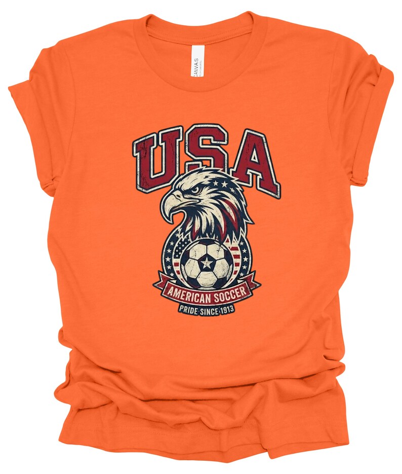 K&ouml;nnte beinhalten: Ein orangefarbenes T-Shirt mit kurzen &Auml;rmeln und einer Grafik eines Wei&szlig;kopfseeadler-Kopfes &uuml;ber einem Fu&szlig;ball. Der Text lautet USA, AMERICAN SOCCER und PRIDE SINCE 1913. Das Design enth&auml;lt rote, wei&szlig;e und blaue Farben mit einer abgenutzten Textur.