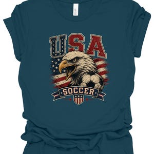 Peut inclure: Un t-shirt &agrave; manches courtes bleu sarcelle fonc&eacute; avec une impression graphique d'un pygargue &agrave; t&ecirc;te blanche, d'un ballon de football et du drapeau am&eacute;ricain. Le texte USA est imprim&eacute; en rouge et bleu au-dessus de l'aigle et SOCCER sur une banni&egrave;re.