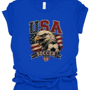 Peut inclure: Un t-shirt bleu roi avec une impression graphique repr&eacute;sentant un pygargue &agrave; t&ecirc;te blanche, un ballon de football et le drapeau am&eacute;ricain. Le texte "USA" appara&icirc;t en grandes lettres rouges et bleues au-dessus de l'aigle, avec "SOCCER" sur une banni&egrave;re.