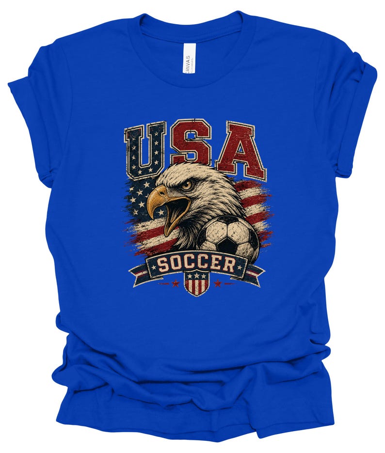 Peut inclure: Un t-shirt bleu roi avec une impression graphique repr&eacute;sentant un pygargue &agrave; t&ecirc;te blanche, un ballon de football et le drapeau am&eacute;ricain. Le texte "USA" appara&icirc;t en grandes lettres rouges et bleues au-dessus de l'aigle, avec "SOCCER" sur une banni&egrave;re.