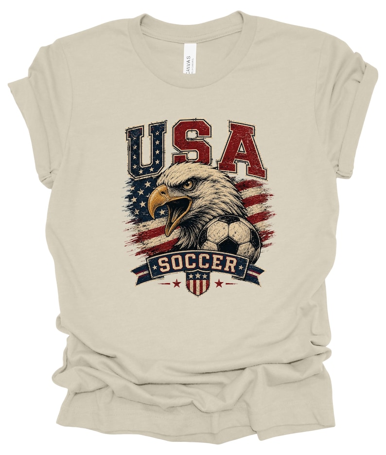 Peut inclure: Un t-shirt beige &agrave; manches courtes avec une impression graphique repr&eacute;sentant un pygargue &agrave; t&ecirc;te blanche, un ballon de football et le drapeau am&eacute;ricain. Le texte "USA" est &eacute;crit au-dessus et "SOCCER" figure sur une banni&egrave;re en dessous.