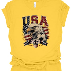 Peut inclure: Un t-shirt jaune vif &agrave; manches courtes avec une impression graphique repr&eacute;sentant un pygargue &agrave; t&ecirc;te blanche, un ballon de football et le drapeau am&eacute;ricain. Le texte "USA" est &eacute;crit en grandes lettres rouges et bleues au-dessus de l'aigle.