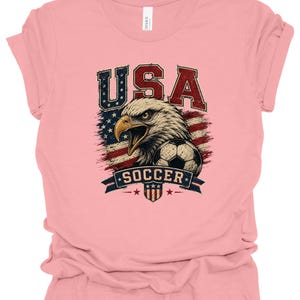 Peut inclure: Un t-shirt &agrave; manches courtes rose clair avec une impression graphique repr&eacute;sentant un pygargue &agrave; t&ecirc;te blanche, un ballon de football et le drapeau am&eacute;ricain. Le texte USA est imprim&eacute; au-dessus de l'aigle et SOCCER sur une banni&egrave;re en dessous.