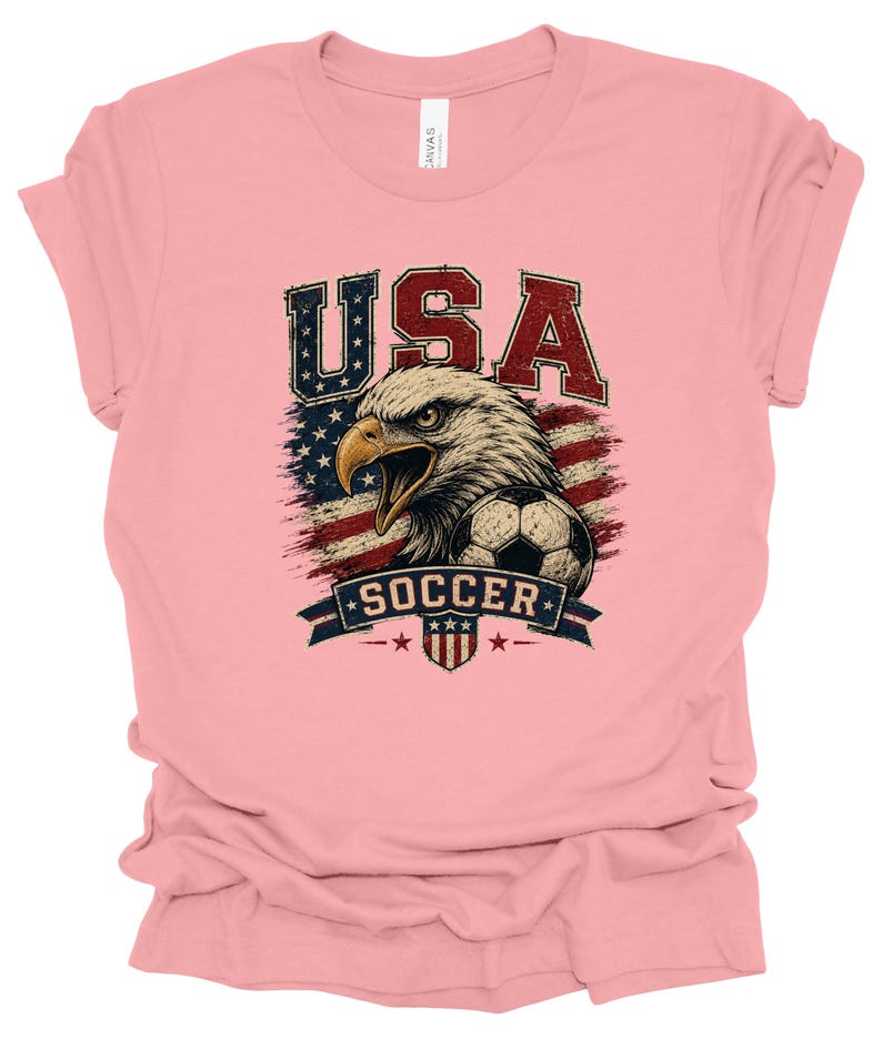Peut inclure: Un t-shirt &agrave; manches courtes rose clair avec une impression graphique repr&eacute;sentant un pygargue &agrave; t&ecirc;te blanche, un ballon de football et le drapeau am&eacute;ricain. Le texte USA est imprim&eacute; au-dessus de l'aigle et SOCCER sur une banni&egrave;re en dessous.
