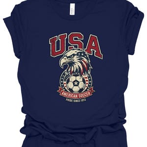 K&ouml;nnte beinhalten: Ein dunkelblaues Kurzarm-T-Shirt mit einer Grafik eines Wei&szlig;kopfseeadler-Kopfes &uuml;ber einem Fu&szlig;ball, umgeben von Sternen und Streifen. Der Text lautet USA, AMERICAN SOCCER und PRIDE SINCE 1913 in einer abgenutzten, roten und wei&szlig;en Schrift.