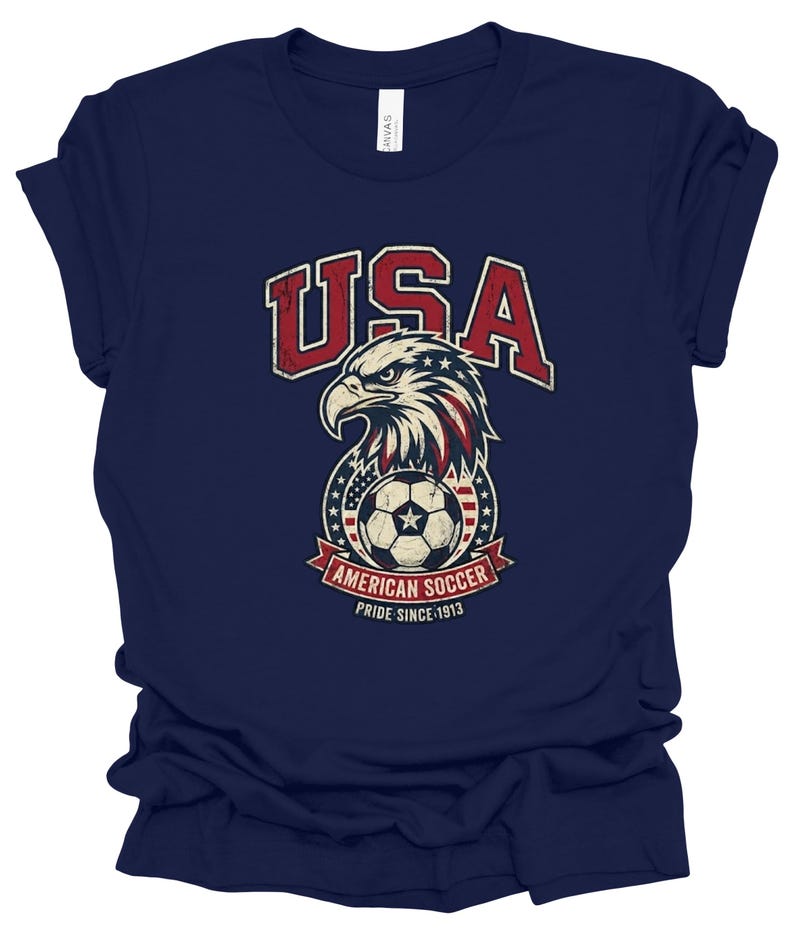 K&ouml;nnte beinhalten: Ein dunkelblaues Kurzarm-T-Shirt mit einer Grafik eines Wei&szlig;kopfseeadler-Kopfes &uuml;ber einem Fu&szlig;ball, umgeben von Sternen und Streifen. Der Text lautet USA, AMERICAN SOCCER und PRIDE SINCE 1913 in einer abgenutzten, roten und wei&szlig;en Schrift.