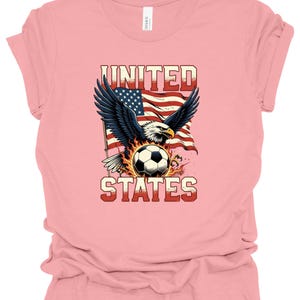 Op de afbeelding: Een lichtroze t-shirt met een afbeelding van een Amerikaanse zeearend met gespreide vleugels boven een voetbal omringd door vlammen. Achter de adelaar wappert een Amerikaanse vlag. De tekst UNITED en STATES is op het shirt gedrukt.