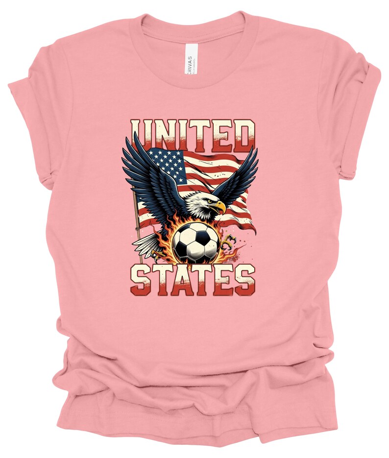 Op de afbeelding: Een lichtroze t-shirt met een afbeelding van een Amerikaanse zeearend met gespreide vleugels boven een voetbal omringd door vlammen. Achter de adelaar wappert een Amerikaanse vlag. De tekst UNITED en STATES is op het shirt gedrukt.