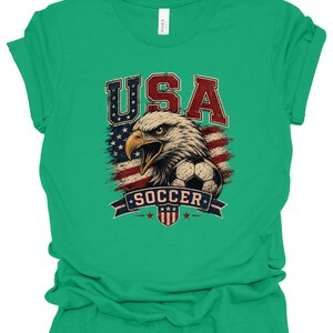 Peut inclure: Un t-shirt vert vif &agrave; manches courtes avec un motif vieilli repr&eacute;sentant un pygargue &agrave; t&ecirc;te blanche, un ballon de football et le drapeau am&eacute;ricain. Le texte USA est en rouge et bleu au-dessus, et le mot SOCCER est sur une banni&egrave;re bleue.