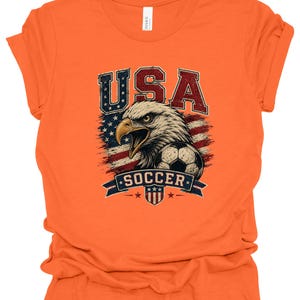 Peut inclure: Un t-shirt orange &agrave; manches courtes avec une impression graphique repr&eacute;sentant un pygargue &agrave; t&ecirc;te blanche, un ballon de football et le drapeau am&eacute;ricain. Le texte USA est imprim&eacute; au-dessus de l'aigle et SOCCER sur une banni&egrave;re bleue.