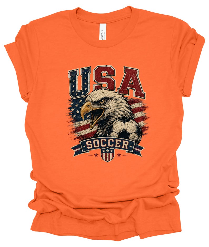 Peut inclure: Un t-shirt orange &agrave; manches courtes avec une impression graphique repr&eacute;sentant un pygargue &agrave; t&ecirc;te blanche, un ballon de football et le drapeau am&eacute;ricain. Le texte USA est imprim&eacute; au-dessus de l'aigle et SOCCER sur une banni&egrave;re bleue.