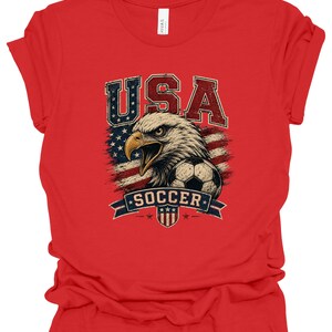 Peut inclure: Un t-shirt rouge &agrave; manches courtes avec une impression graphique repr&eacute;sentant un pygargue &agrave; t&ecirc;te blanche, un ballon de football et un drapeau am&eacute;ricain stylis&eacute;. Le texte USA est imprim&eacute; au-dessus de l'aigle et SOCCER sur une banni&egrave;re bleue.