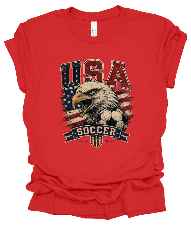 Peut inclure: Un t-shirt rouge &agrave; manches courtes avec une impression graphique repr&eacute;sentant un pygargue &agrave; t&ecirc;te blanche, un ballon de football et un drapeau am&eacute;ricain stylis&eacute;. Le texte USA est imprim&eacute; au-dessus de l'aigle et SOCCER sur une banni&egrave;re bleue.