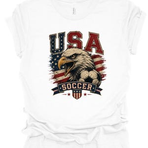 Peut inclure: Un t-shirt blanc &agrave; manches courtes avec une impression graphique repr&eacute;sentant un pygargue &agrave; t&ecirc;te blanche, un ballon de football et le drapeau am&eacute;ricain. Le texte USA est imprim&eacute; en grandes lettres au-dessus de l'aigle et SOCCER sur une banni&egrave;re.