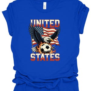 Op de afbeelding: Een koningsblauw t-shirt met korte mouwen en een grafische print van een Amerikaanse zeearend met gespreide vleugels boven een voetbal omringd door vlammen. Achter de adelaar wappert een Amerikaanse vlag. De tekst UNITED staat boven en STATES onder.