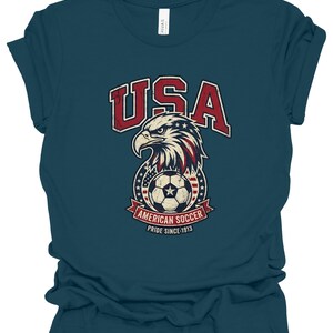 K&ouml;nnte beinhalten: Ein dunkelblaugr&uuml;nes Kurzarm-T-Shirt mit einer abgenutzten Grafik eines Wei&szlig;kopfseeadler-Kopfes &uuml;ber einem Fu&szlig;ball, umgeben von Sternen und Streifen. Der Text lautet USA, AMERICAN SOCCER und PRIDE SINCE 1913 in Rot, Wei&szlig; und Blau.