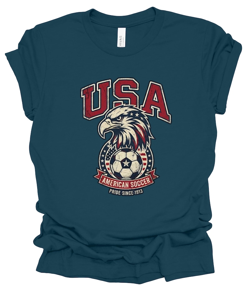 K&ouml;nnte beinhalten: Ein dunkelblaugr&uuml;nes Kurzarm-T-Shirt mit einer abgenutzten Grafik eines Wei&szlig;kopfseeadler-Kopfes &uuml;ber einem Fu&szlig;ball, umgeben von Sternen und Streifen. Der Text lautet USA, AMERICAN SOCCER und PRIDE SINCE 1913 in Rot, Wei&szlig; und Blau.