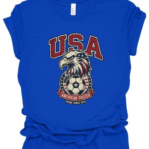 K&ouml;nnte beinhalten: Ein k&ouml;nigsblaues Kurzarm-T-Shirt mit einem Grafikdruck eines Wei&szlig;kopfseeadler-Kopfes &uuml;ber einem Fu&szlig;ball. Das Design enth&auml;lt den Text USA, AMERICAN SOCCER und PRIDE SINCE 1913 in Rot, Wei&szlig; und Blau mit einer abgenutzten, vintage-artigen Textur.