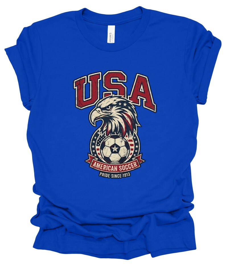 K&ouml;nnte beinhalten: Ein k&ouml;nigsblaues Kurzarm-T-Shirt mit einem Grafikdruck eines Wei&szlig;kopfseeadler-Kopfes &uuml;ber einem Fu&szlig;ball. Das Design enth&auml;lt den Text USA, AMERICAN SOCCER und PRIDE SINCE 1913 in Rot, Wei&szlig; und Blau mit einer abgenutzten, vintage-artigen Textur.