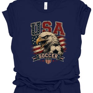 Peut inclure: Un t-shirt bleu marine avec une impression graphique repr&eacute;sentant un pygargue &agrave; t&ecirc;te blanche, un ballon de football et un drapeau am&eacute;ricain vieilli. Le texte USA est imprim&eacute; en rouge et bleu au-dessus, et SOCCER figure sur une banni&egrave;re.