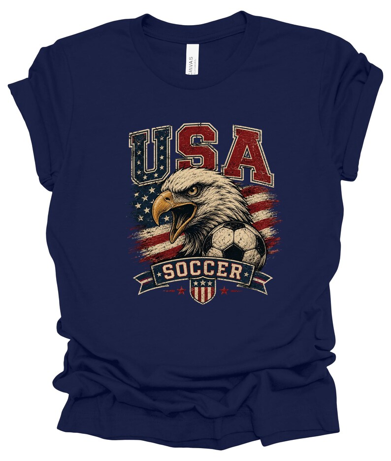 Peut inclure: Un t-shirt bleu marine avec une impression graphique repr&eacute;sentant un pygargue &agrave; t&ecirc;te blanche, un ballon de football et un drapeau am&eacute;ricain vieilli. Le texte USA est imprim&eacute; en rouge et bleu au-dessus, et SOCCER figure sur une banni&egrave;re.