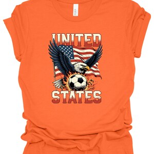 Op de afbeelding: Een oranje T-shirt met korte mouwen en een afbeelding van een Amerikaanse zeearend met gespreide vleugels boven een voetbal, tegen een wapperende Amerikaanse vlag. De tekst UNITED staat in het rood erboven en STATES in het rood eronder.