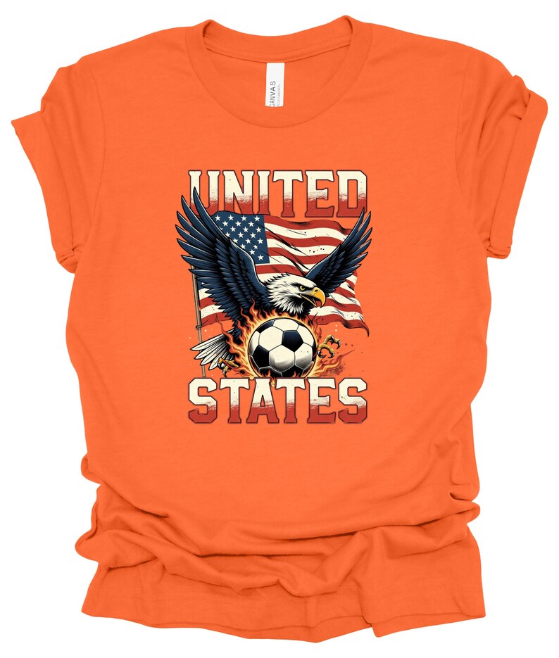 Op de afbeelding: Een oranje T-shirt met korte mouwen en een afbeelding van een Amerikaanse zeearend met gespreide vleugels boven een voetbal, tegen een wapperende Amerikaanse vlag. De tekst UNITED staat in het rood erboven en STATES in het rood eronder.