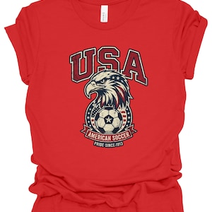 K&ouml;nnte beinhalten: Ein rotes Kurzarm-T-Shirt mit einem Grafikdruck eines Wei&szlig;kopfseeadler-Kopfes &uuml;ber einem Fu&szlig;ball. Der Text lautet USA, AMERICAN SOCCER und PRIDE SINCE 1913. Das Design enth&auml;lt rote, wei&szlig;e und blaue Farben mit einer abgenutzten Textur.