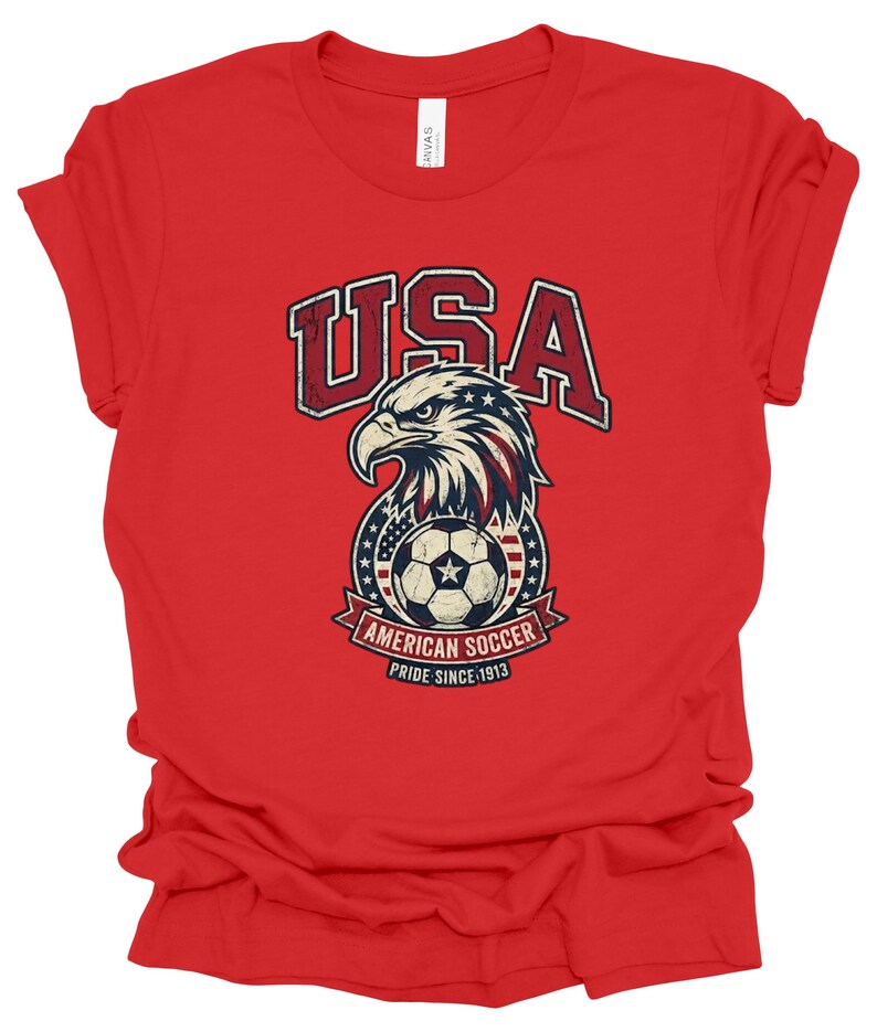K&ouml;nnte beinhalten: Ein rotes Kurzarm-T-Shirt mit einem Grafikdruck eines Wei&szlig;kopfseeadler-Kopfes &uuml;ber einem Fu&szlig;ball. Der Text lautet USA, AMERICAN SOCCER und PRIDE SINCE 1913. Das Design enth&auml;lt rote, wei&szlig;e und blaue Farben mit einer abgenutzten Textur.