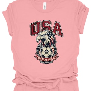 K&ouml;nnte beinhalten: Ein hellrosa T-Shirt mit kurzen &Auml;rmeln und einer Grafik eines Wei&szlig;kopfseeadler-Kopfes &uuml;ber einem Fu&szlig;ball. Der Text lautet USA, AMERICAN SOCCER und PRIDE SINCE 1913. Das Design enth&auml;lt rote, wei&szlig;e und blaue Farben mit einer abgenutzten Textur.