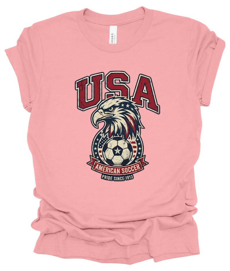 K&ouml;nnte beinhalten: Ein hellrosa T-Shirt mit kurzen &Auml;rmeln und einer Grafik eines Wei&szlig;kopfseeadler-Kopfes &uuml;ber einem Fu&szlig;ball. Der Text lautet USA, AMERICAN SOCCER und PRIDE SINCE 1913. Das Design enth&auml;lt rote, wei&szlig;e und blaue Farben mit einer abgenutzten Textur.