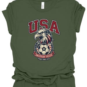 K&ouml;nnte beinhalten: Ein olivgr&uuml;nes Kurzarm-T-Shirt mit einer Grafik eines Wei&szlig;kopfseeadler-Kopfes &uuml;ber einem Fu&szlig;ball. Das Design enth&auml;lt den Text USA, AMERICAN SOCCER und PRIDE SINCE 1913 in Rot, Wei&szlig; und Blau mit einer abgenutzten, vintage-artigen Textur.