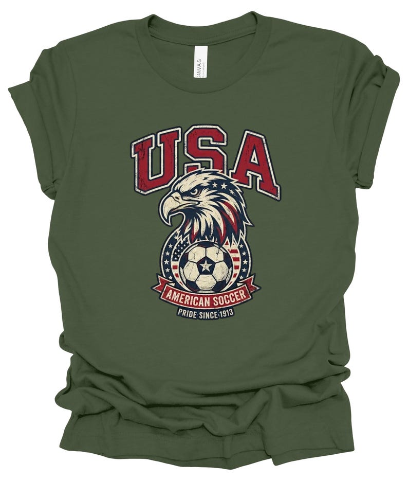 K&ouml;nnte beinhalten: Ein olivgr&uuml;nes Kurzarm-T-Shirt mit einer Grafik eines Wei&szlig;kopfseeadler-Kopfes &uuml;ber einem Fu&szlig;ball. Das Design enth&auml;lt den Text USA, AMERICAN SOCCER und PRIDE SINCE 1913 in Rot, Wei&szlig; und Blau mit einer abgenutzten, vintage-artigen Textur.