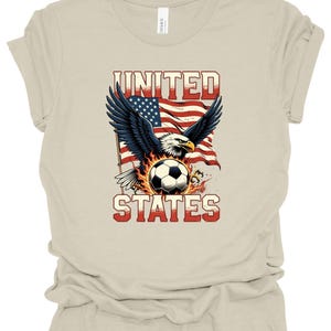 Op de afbeelding: Een beige T-shirt met korte mouwen en een afbeelding van een Amerikaanse zeearend met gespreide vleugels boven een voetbal omringd door vlammen. Achter de adelaar staat een Amerikaanse vlag. De tekst UNITED en STATES is op het shirt gedrukt.