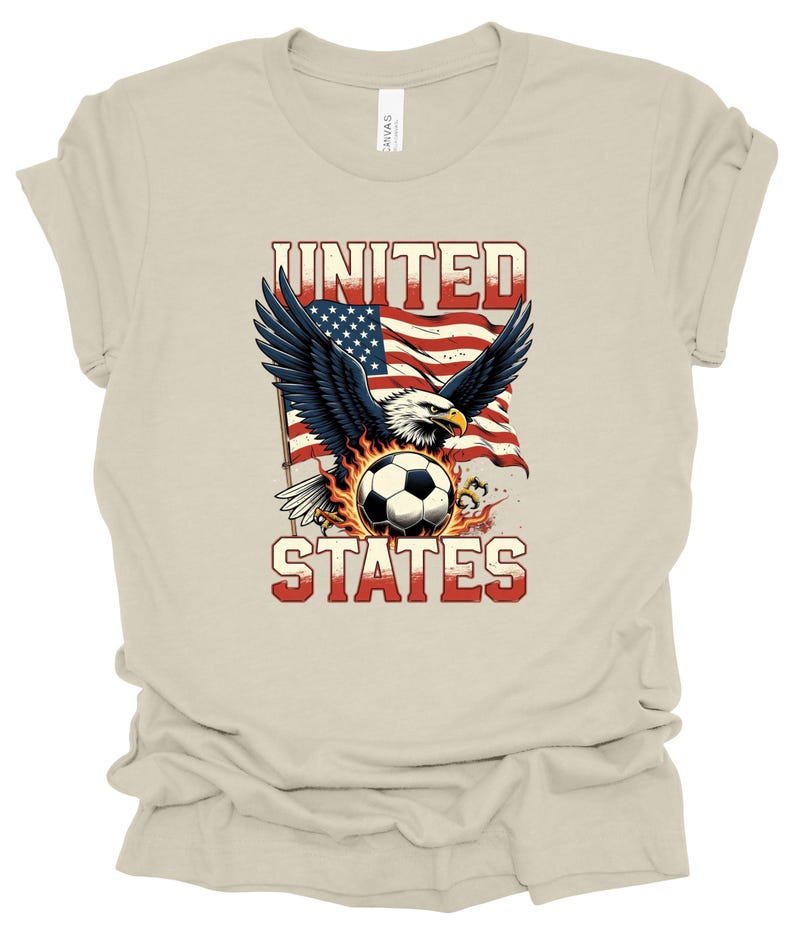 Op de afbeelding: Een beige T-shirt met korte mouwen en een afbeelding van een Amerikaanse zeearend met gespreide vleugels boven een voetbal omringd door vlammen. Achter de adelaar staat een Amerikaanse vlag. De tekst UNITED en STATES is op het shirt gedrukt.