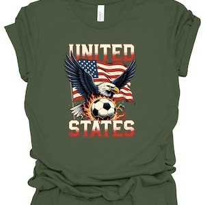 Op de afbeelding: Een olijfgroen t-shirt met een afbeelding van een Amerikaanse zeearend met gespreide vleugels boven een voetbal omringd door vlammen. Achter de adelaar is een Amerikaanse vlag te zien. De tekst UNITED staat boven de adelaar en STATES eronder.