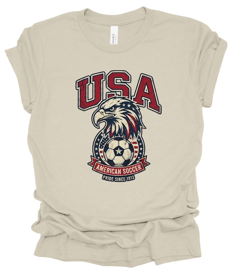 K&ouml;nnte beinhalten: Ein beiges Kurzarm-T-Shirt mit einer Grafik eines Wei&szlig;kopfseeadler-Kopfes &uuml;ber einem Fu&szlig;ball. Das Design enth&auml;lt den Text USA, AMERICAN SOCCER und PRIDE SINCE 1913 in den Farben Rot, Wei&szlig; und Blau mit einer abgenutzten, vintage-artigen Textur.