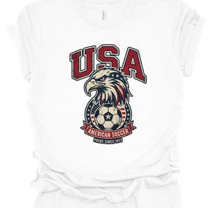 K&ouml;nnte beinhalten: Ein wei&szlig;es kurz&auml;rmeliges T-Shirt mit einer Grafik eines Wei&szlig;kopfseeadler-Kopfes &uuml;ber einem Fu&szlig;ball. Der Text lautet USA, AMERICAN SOCCER und PRIDE SINCE 1913. Das Design enth&auml;lt rote, wei&szlig;e und blaue Farben mit einer abgenutzten Vintage-Textur.