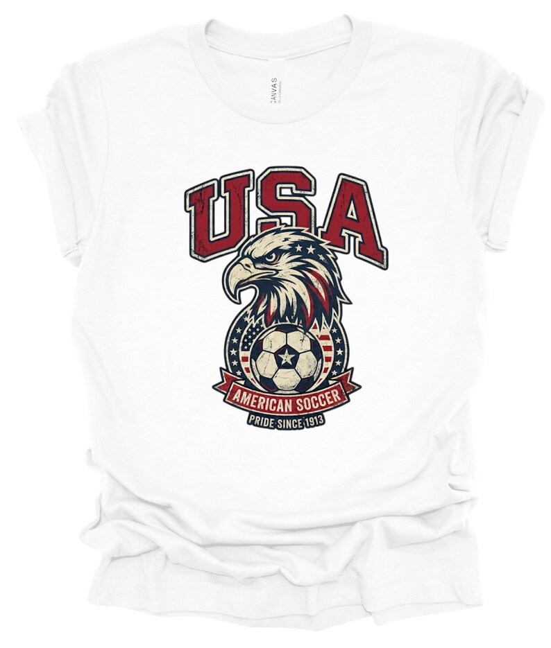 K&ouml;nnte beinhalten: Ein wei&szlig;es kurz&auml;rmeliges T-Shirt mit einer Grafik eines Wei&szlig;kopfseeadler-Kopfes &uuml;ber einem Fu&szlig;ball. Der Text lautet USA, AMERICAN SOCCER und PRIDE SINCE 1913. Das Design enth&auml;lt rote, wei&szlig;e und blaue Farben mit einer abgenutzten Vintage-Textur.