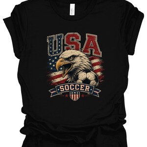 Peut inclure: Un t-shirt noir &agrave; manches courtes avec une impression graphique repr&eacute;sentant un pygargue &agrave; t&ecirc;te blanche, un ballon de football et le drapeau am&eacute;ricain. Le texte USA est imprim&eacute; en grandes lettres au-dessus de l'aigle et SOCCER sur une banni&egrave;re.